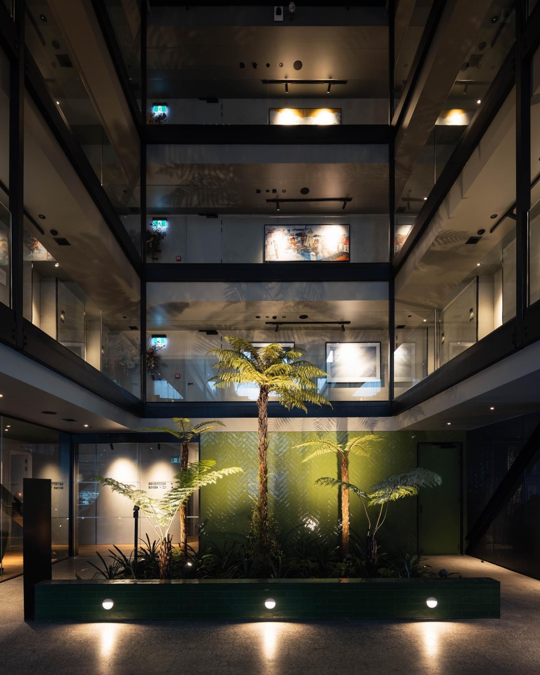 A dimly lit hotel atrium with ferns on the ground floor and artwork displays visible on the 3 floors above it.