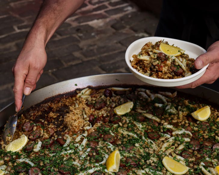 A close up image that features a paella in a large pan, and a man's hand scooping up some of the paella into his own plate.