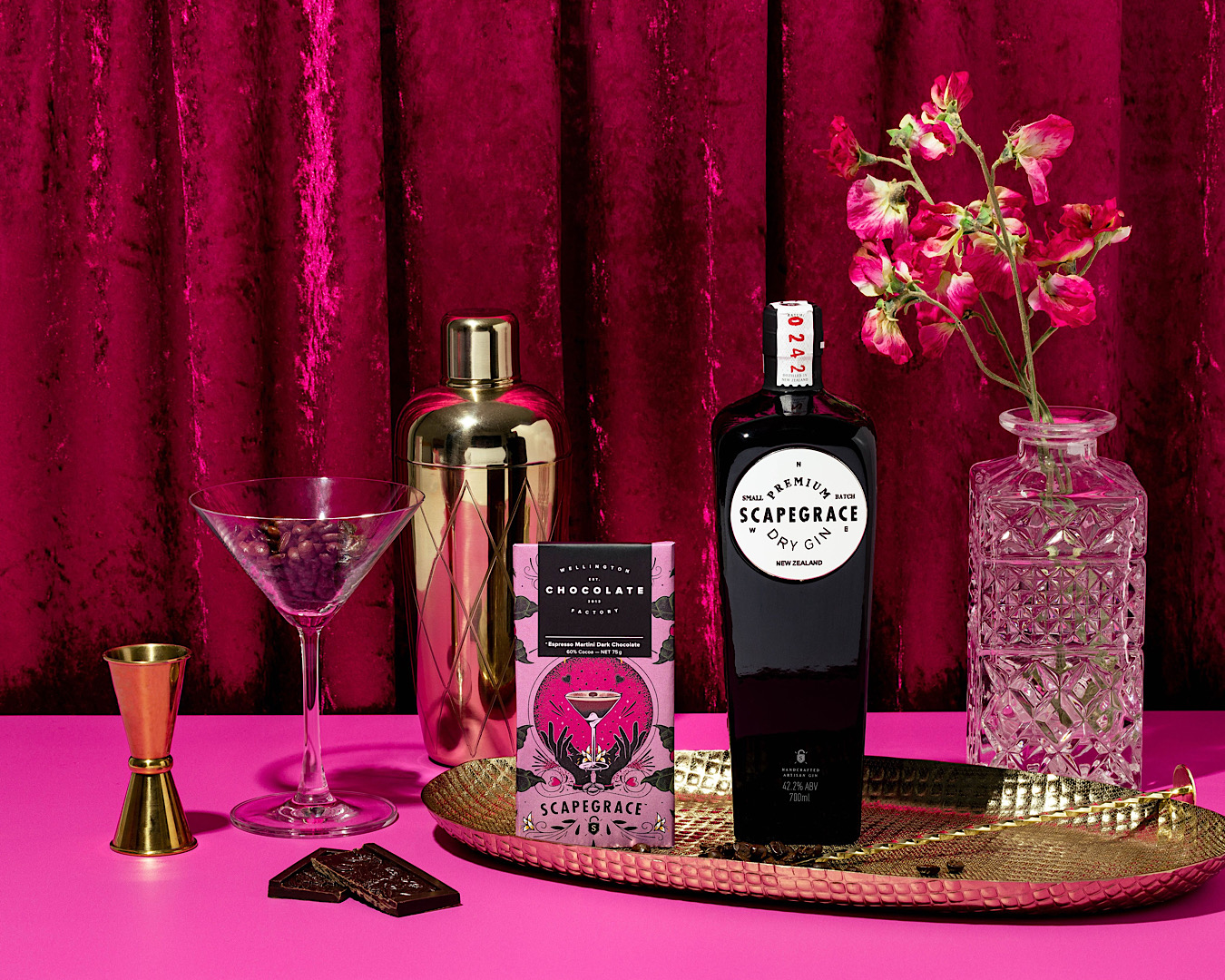 A chocolate bar covered in a pink wrapper of a chic cartoon martini image stands against a pinky red velvet vibes curtain, perched on a gold tray and accompanied by a bottle of Scapegrace gin, a retro vase with flowers, a cocktail shaker and a martini glass filled with coffee beans. 