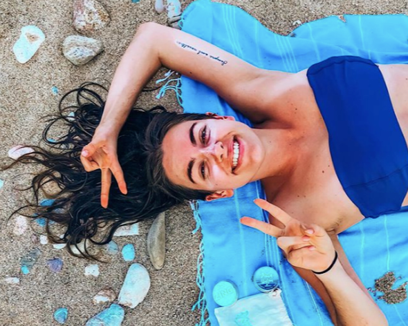 A happy womans makes peace signs with her hands as she lies on the sand wearing a blue bandeau by Salt, one of the coolest swimwear labels in New Zealand. 