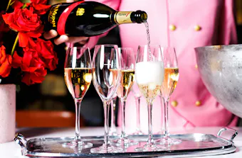 A person in a pink suit pours champagne into flutes on a silver tray.