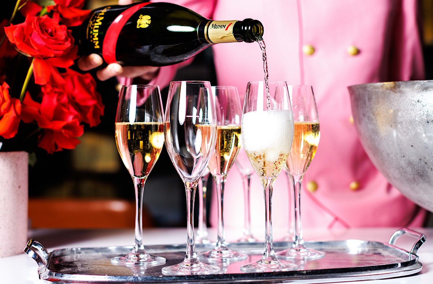 A person in a pink suit pours champagne into flutes on a silver tray.