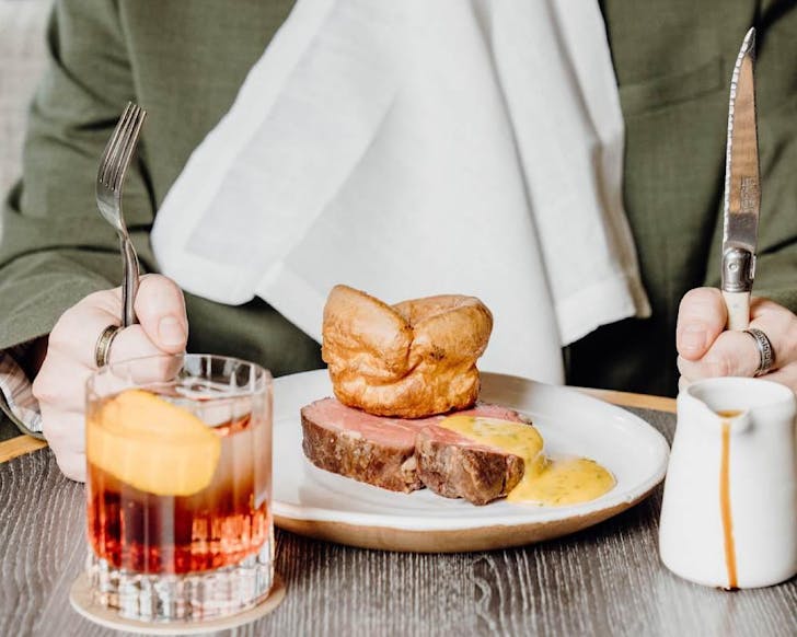 Somebody holds their knife and fork at the ready, about to dive into a slab of stake topped with yellow sauce and a large Yorkshire pudding at Esther Restaurant, one of the best places to go this Fatherâs Day. 