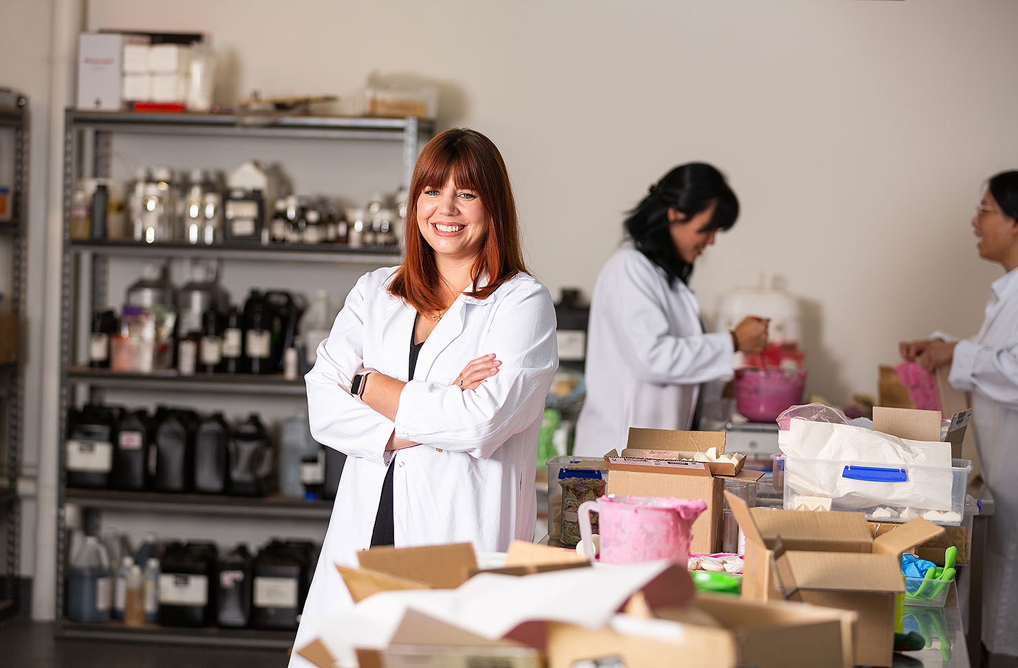 Brianne wearing a white lab coat in Ethique's manufacturing space, with two women making soap mix in the background.