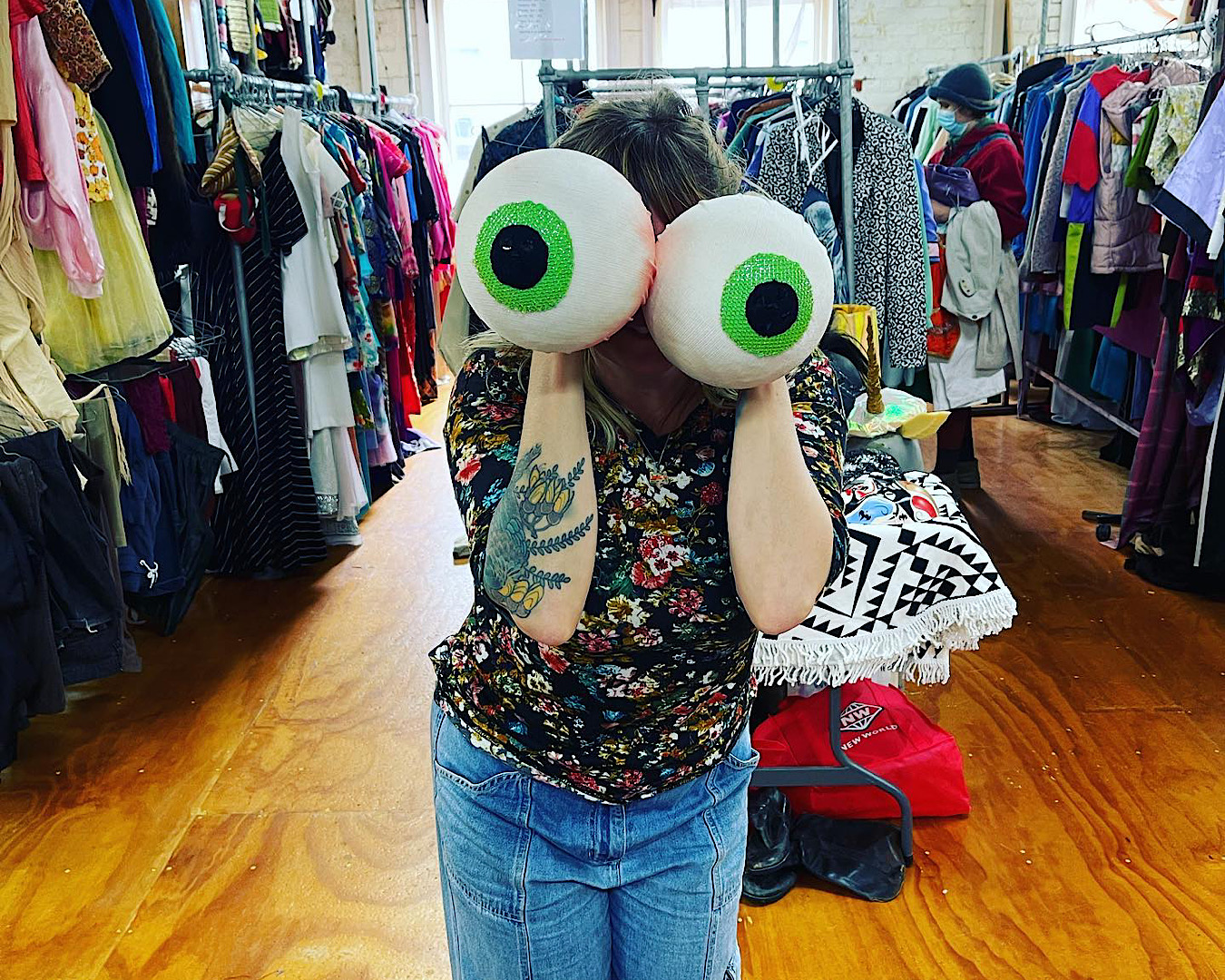 A person in a floral shirt with a kowhai tattoo stands amongst Wellington costume shop The Costume Cave’s racks of clothing holding two giant green eyeballs up to their face.  