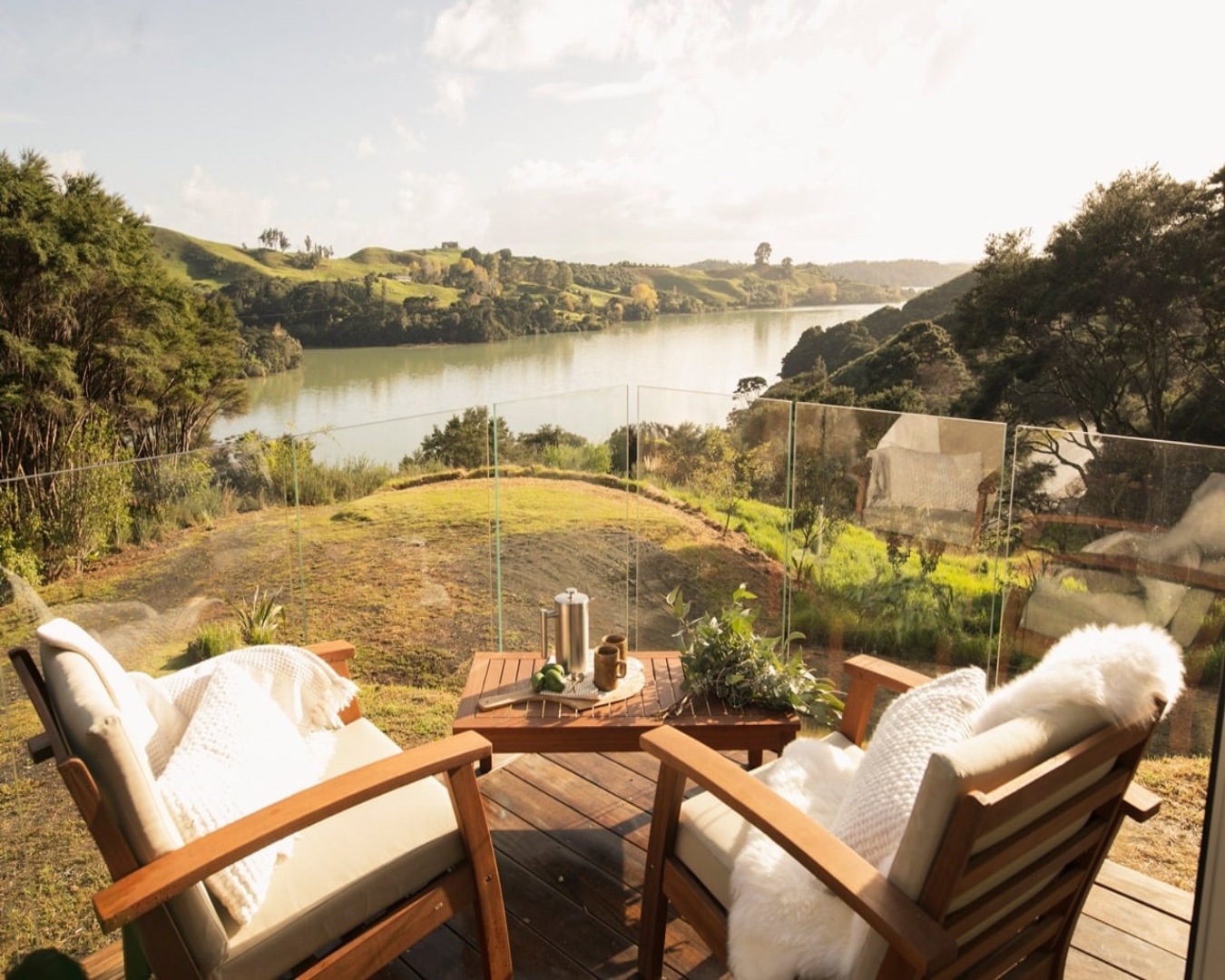 Two cosy chairs at Tarata eco retreat and a coffee pot on a deck overlooking a peaceful-looking harbour.