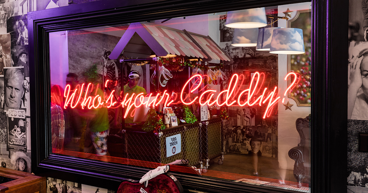 Red neon lettering on a mirror at Holey Moley mini-golf that asks ‘Who’s your caddy?”