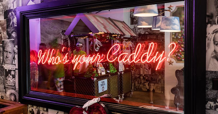 Red neon lettering on a mirror at Holey Moley mini-golf that asks âWhoâs your caddy?â
