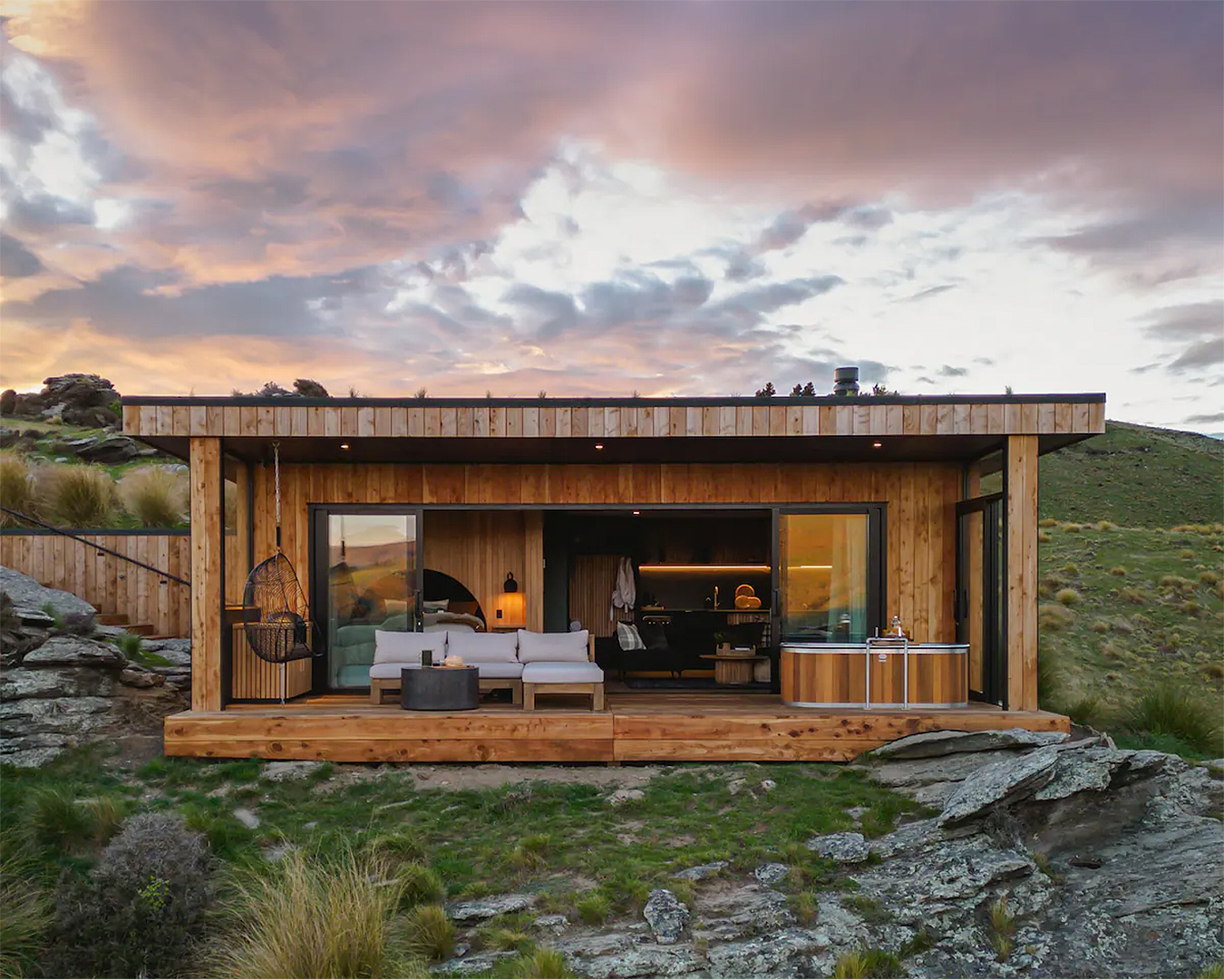 A rectangular, wooden retreat house set on rocks and surrounded by green hills.