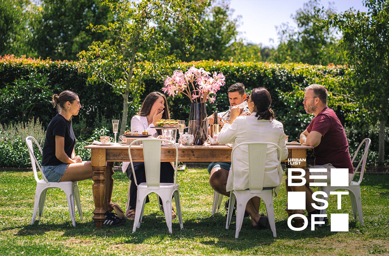 A group of people sit around a high tea table setting in a garden. 