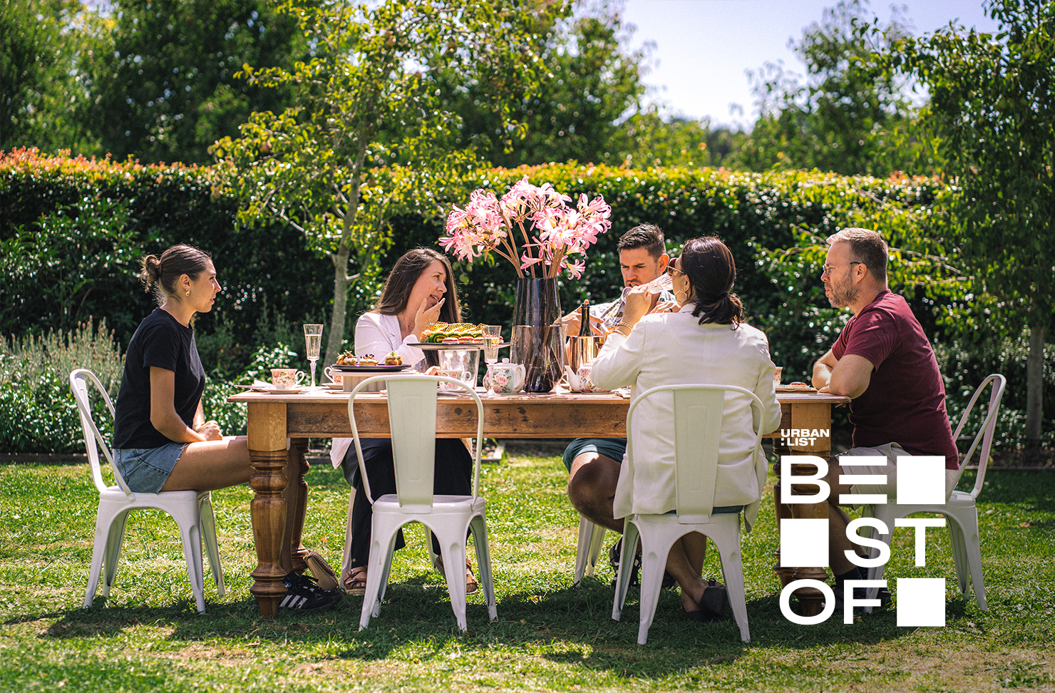 A group of people sit around a high tea table setting in a garden. 