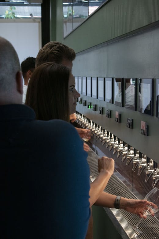 A sage green beer wall with numerous taps lining it's centre on the right hands side of the photograph. Above are slots to place pre-paid cards. On the left hands side, guests are pictured pouring pints of beer into clear glasses.