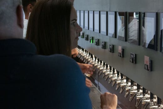 A sage green beer wall with numerous taps lining it's centre on the right hands side of the photograph. Above are slots to place pre-paid cards. On the left hands side, guests are pictured pouring pints of beer into clear glasses.