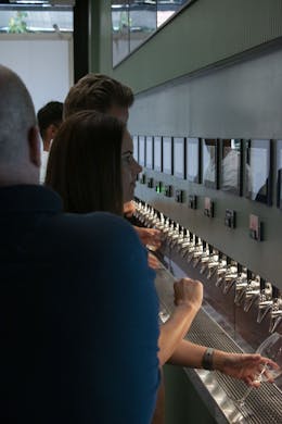 A sage green beer wall with numerous taps lining it's centre on the right hands side of the photograph. Above are slots to place pre-paid cards. On the left hands side, guests are pictured pouring pints of beer into clear glasses.