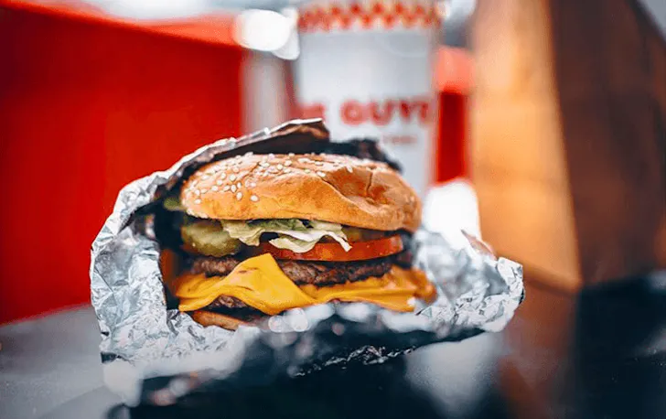 A burger wrapped in foil with a drink in the back. 