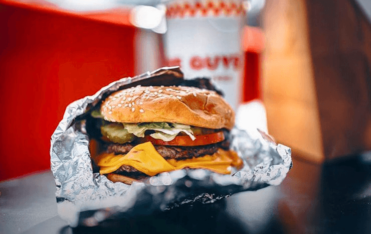 A burger wrapped in foil with a drink in the back. 