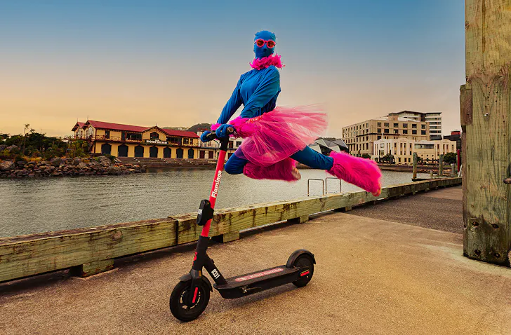 A person dressed in pink and blue leaps on the back of a scooter, cutting a dramatic figure.