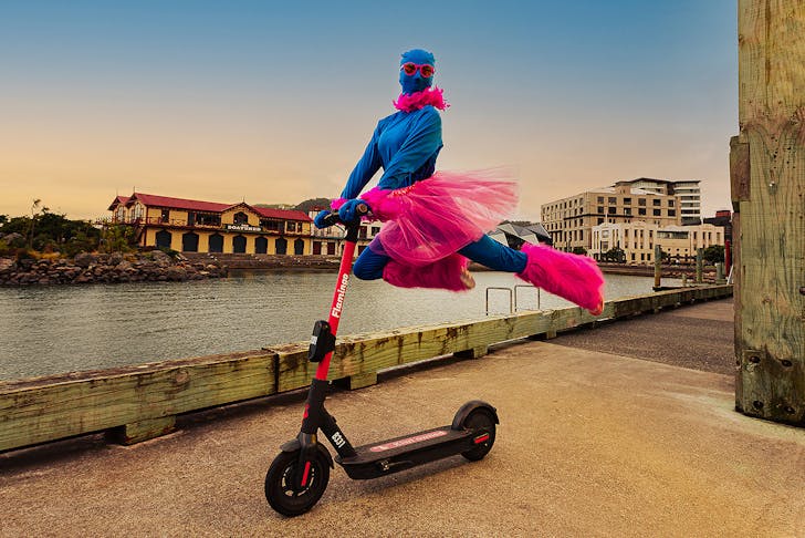 A person dressed in pink and blue leaps on the back of a scooter, cutting a dramatic figure.