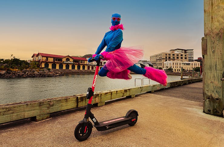 A person dressed in pink and blue leaps on the back of a scooter, cutting a dramatic figure.