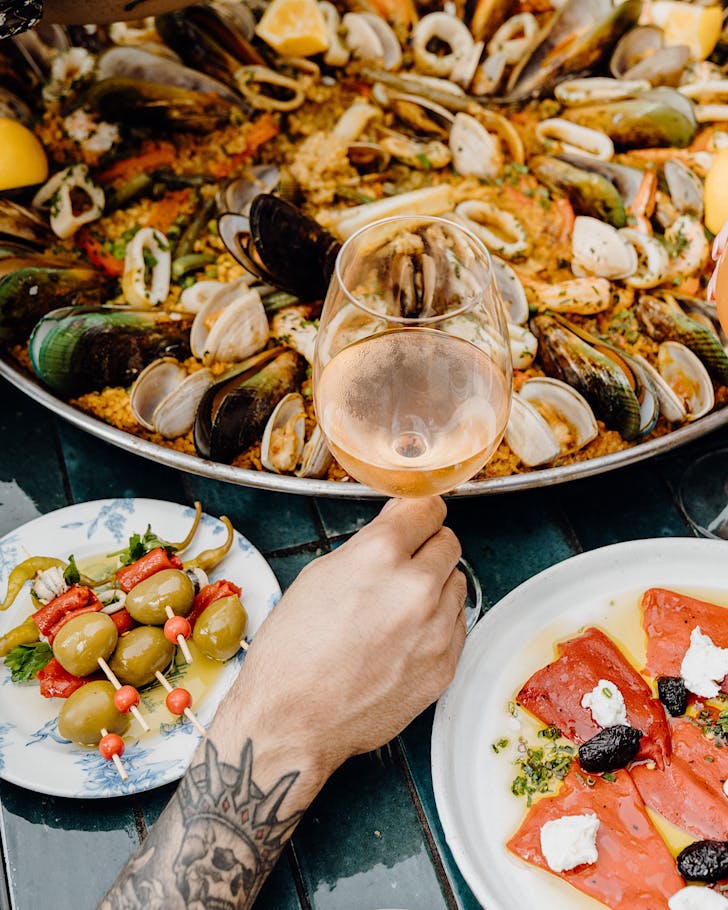 A tattooed forearm holds a glass of white wine in front of a large paella pan and a plate of skewered olives. 