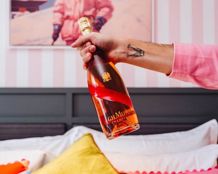 An arm with a champagne flute tattoo holds up a pretty bottle of G.H. Mumm Champagne Cordon Rose. 