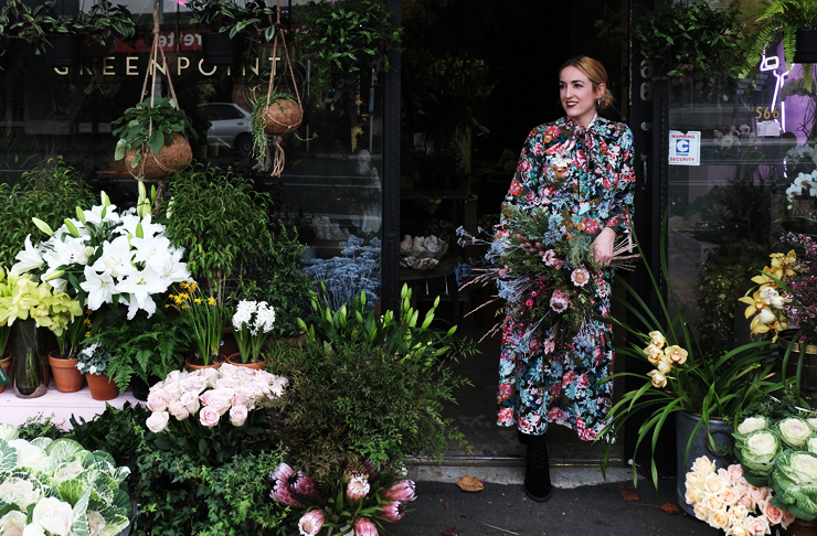 Georgie stands in front of her flower shop, Greenpoint surrounding by flowers and plants. The building is green.