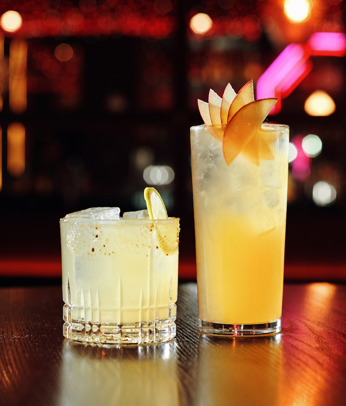 Two yellow-hued cocktails are lined up on the bar. 