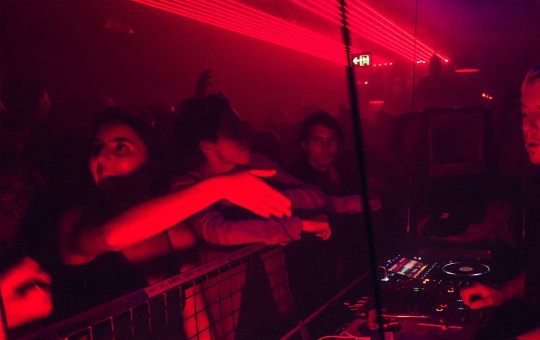 A red-lit nightclub with people dancing and DJ behind the decks.