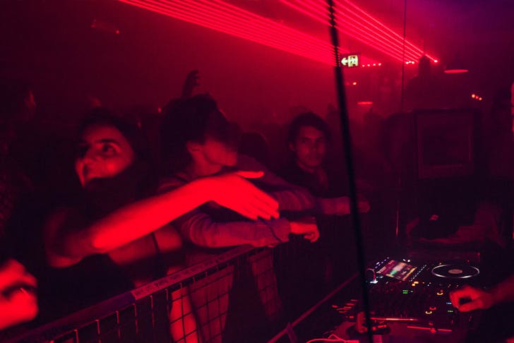 A red-lit nightclub with people dancing and DJ behind the decks.