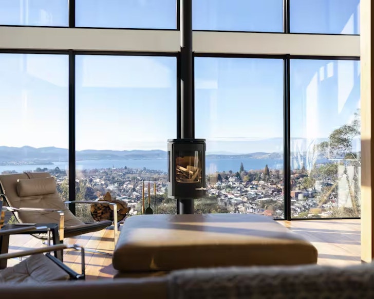 A view of the indoor fireplace in front of the huge window with city views at Glass Holme one of the best Airbnbs in Hobart