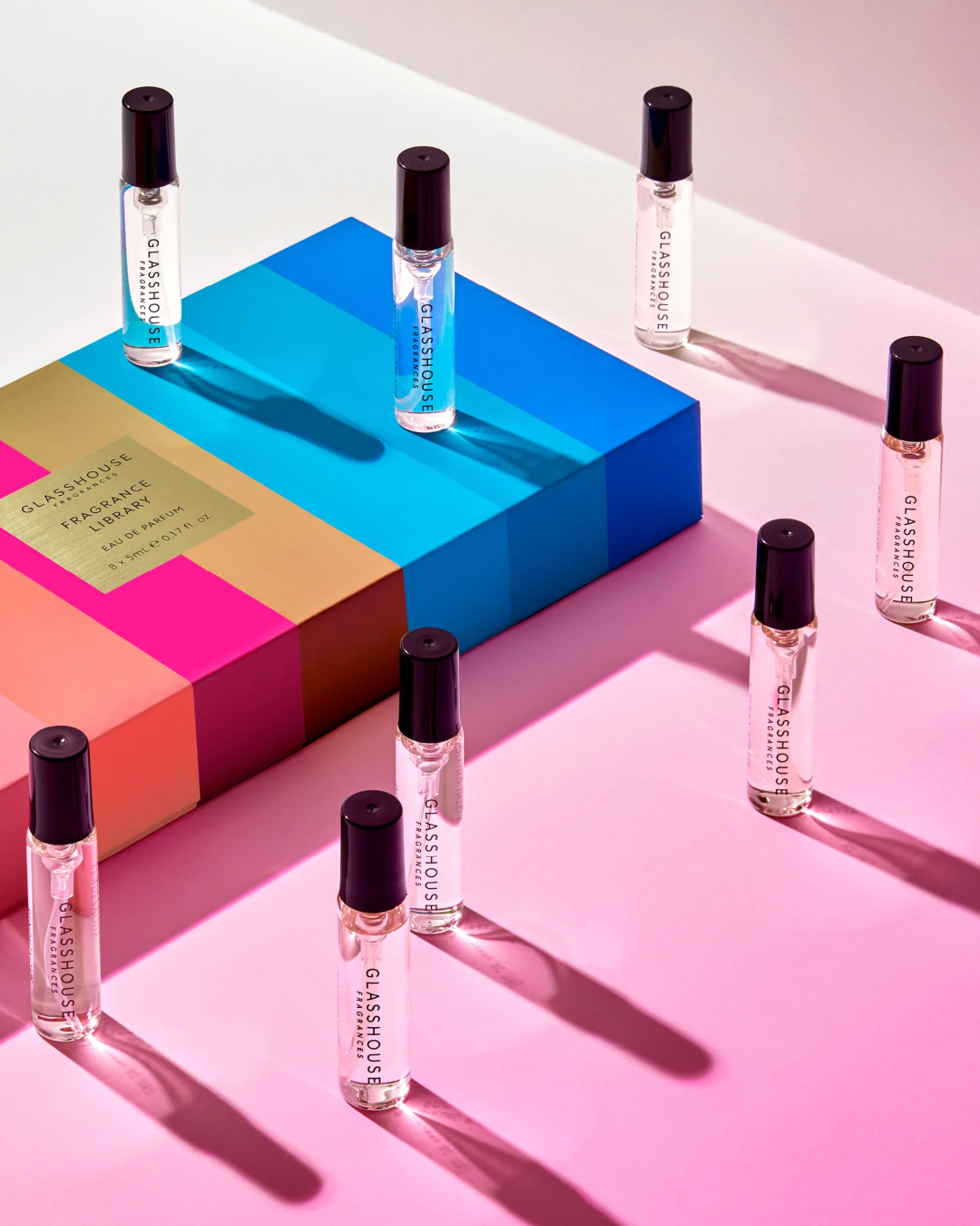 8 small vials of perfume and a colourful striped gift box against a pink backdrop.