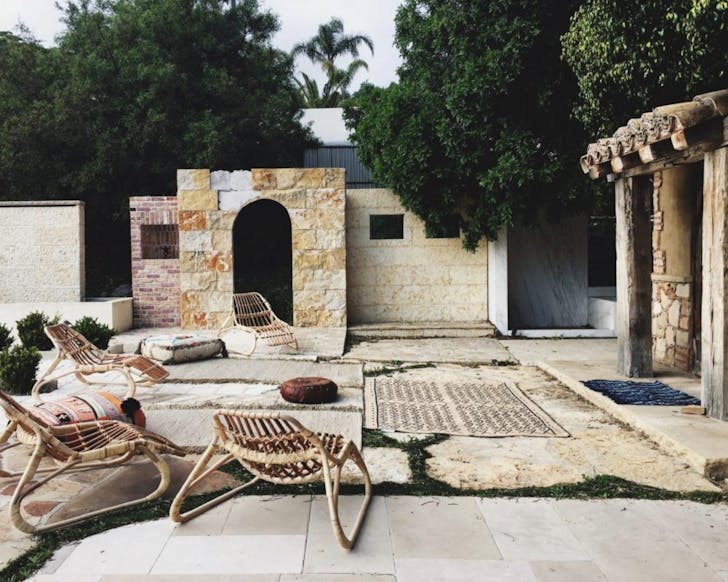 Outdoor setting with wooden lay-back chairs in the forefront, positioned on large sandstone-looking tiles. There is a hut-looking building to the right of the image and giant green trees in the background.