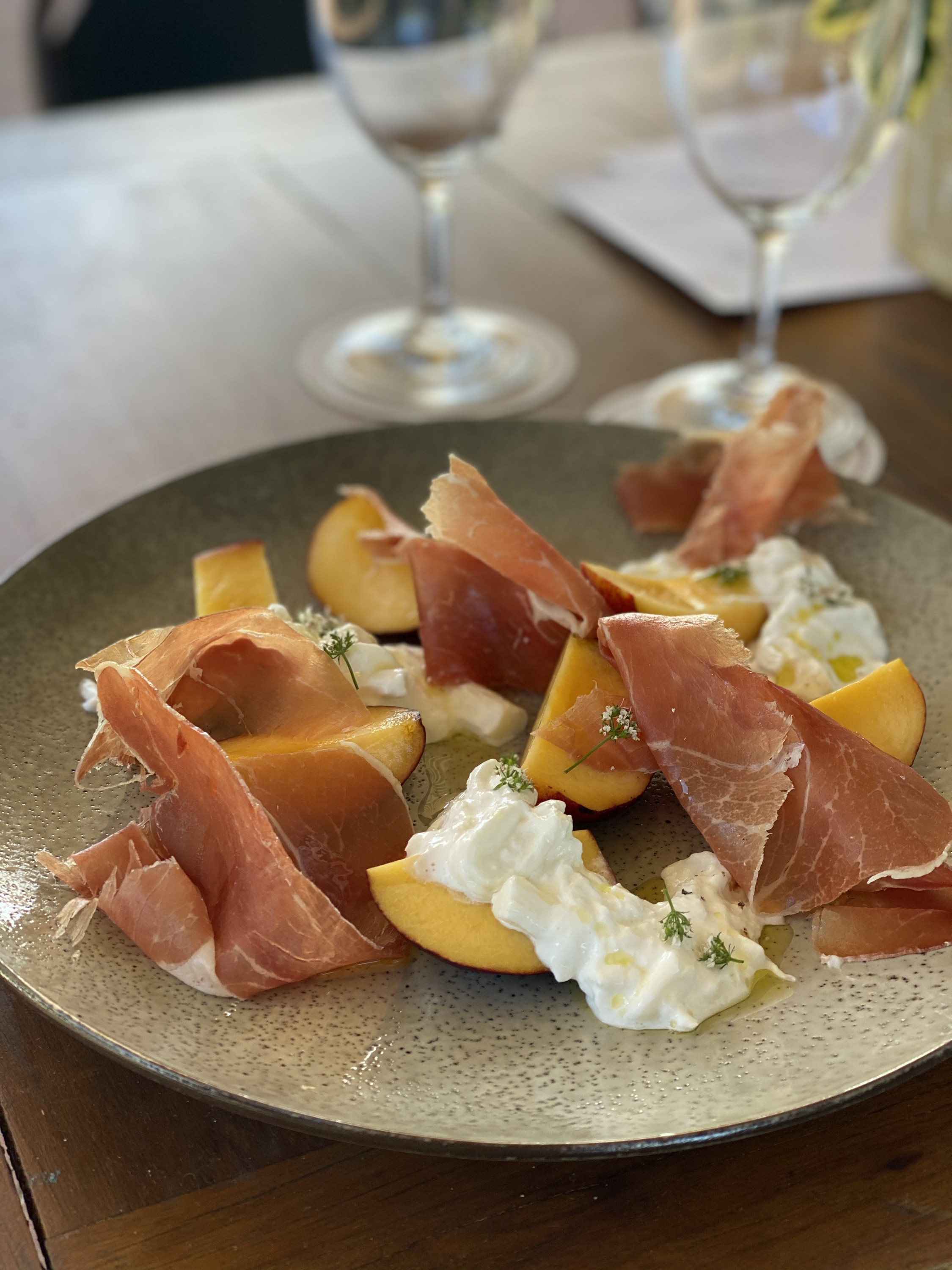 a peach and prosciutto dish with white cheese and a glass of wine in the background