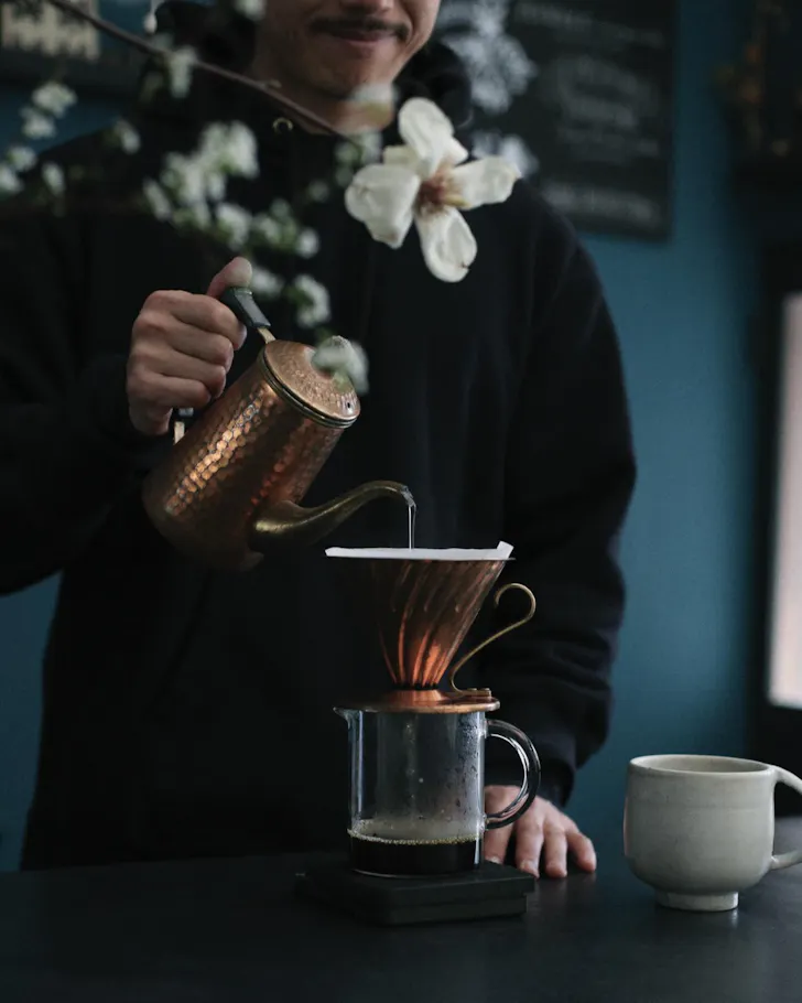 A moody image of a roaster pouring drip coffee with a white flower in the foreground.