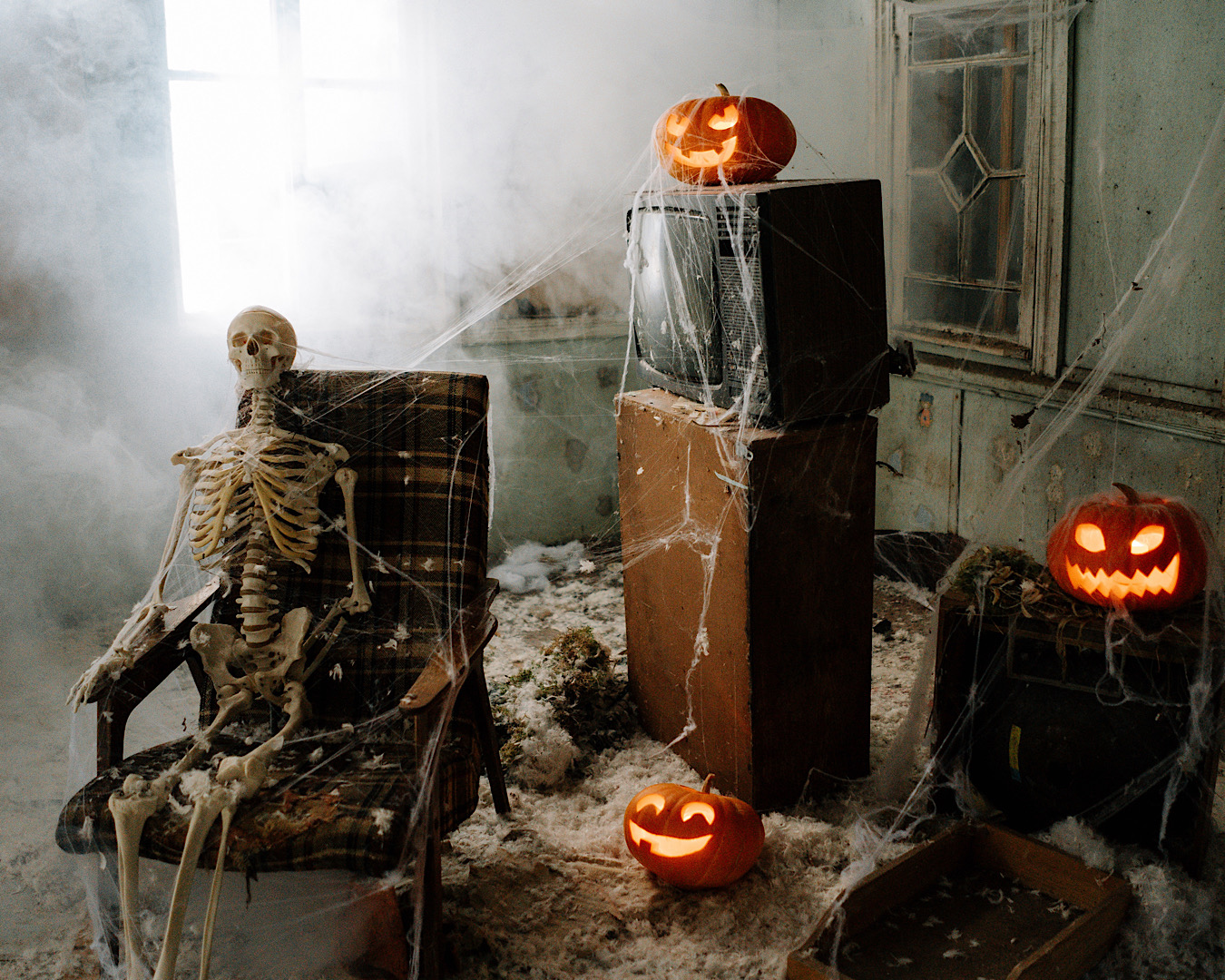 A skeleton sits on a chair in a room covered in cobwebs, peppered with foreboding Jack o’ Lanterns. 