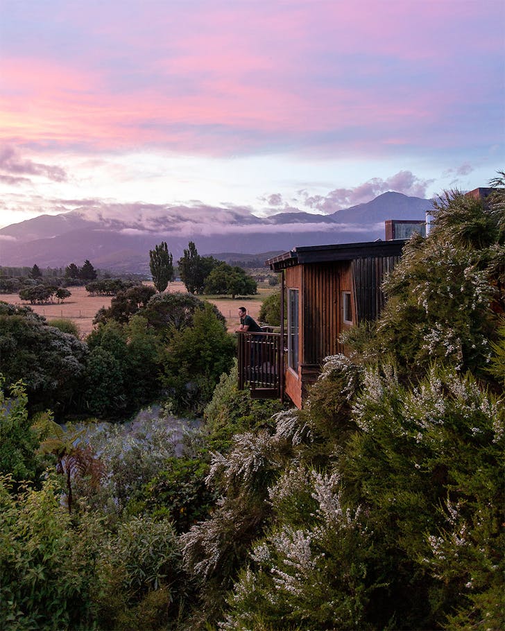 Someone gazes out at the view with mountains behind from a treehouse above the trees.