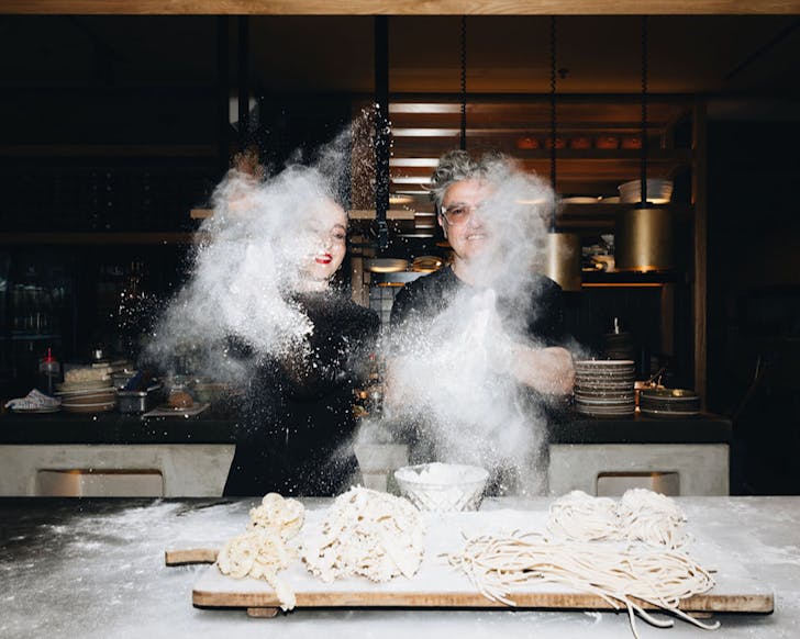 Two people clap floury hands creating a dramatic flour cloud while making bread.