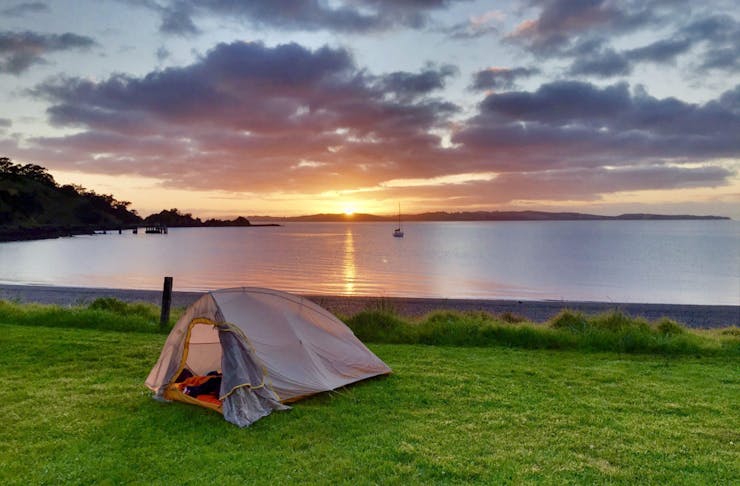 A tent pitched on a grassy beachfront with the sunset on the horizon.