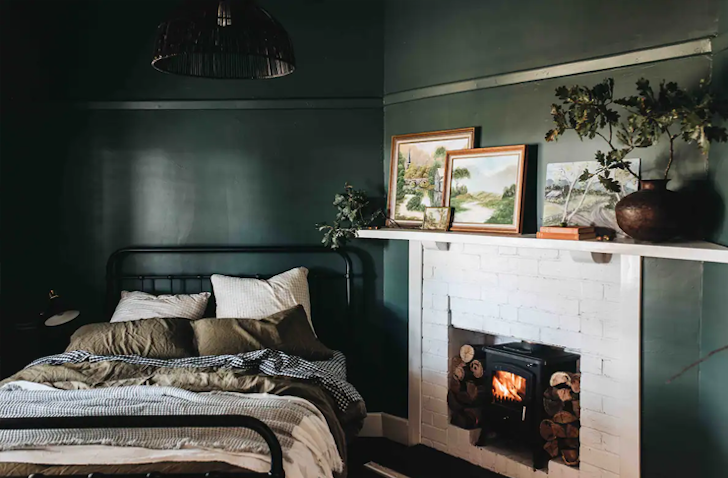 A double sized bed sits in a small emerald green bedroom with a fireplace framed with white brick.