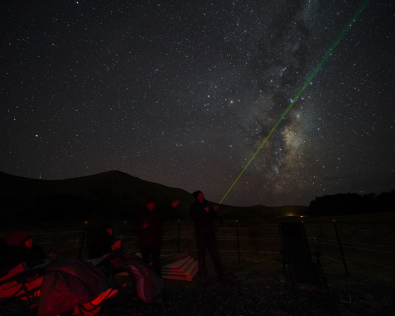 A man points out stars with a laser pointer with people sitting in chairs around nicely rugged up.