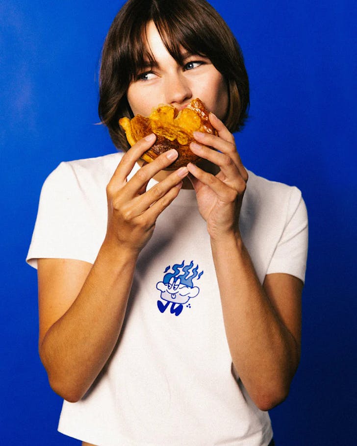 A woman wears a baby tee featuring a mini pie chalk while she eats a Pie Rollas pie. 