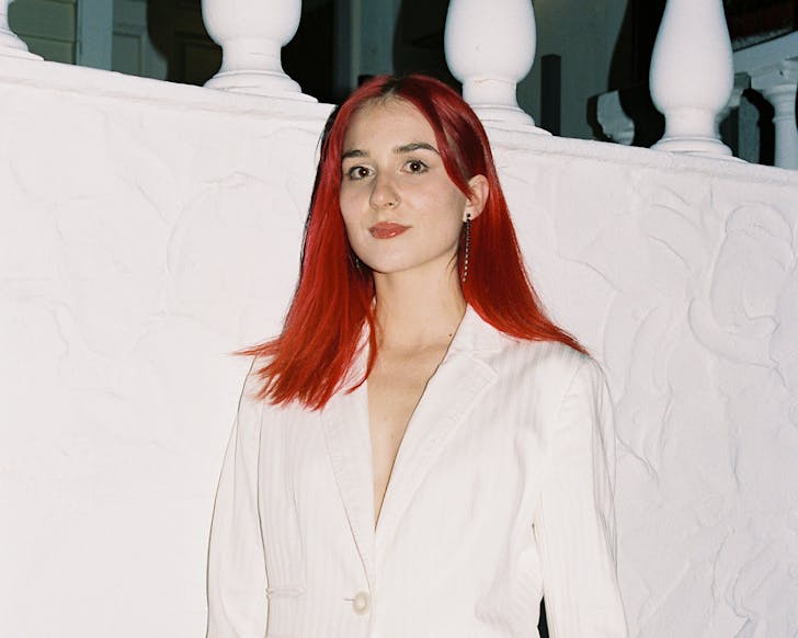 Up and coming DJ Hurricane Emily stands on a white background and looks directly into the camera, her white suit complimenting her red hair. 
