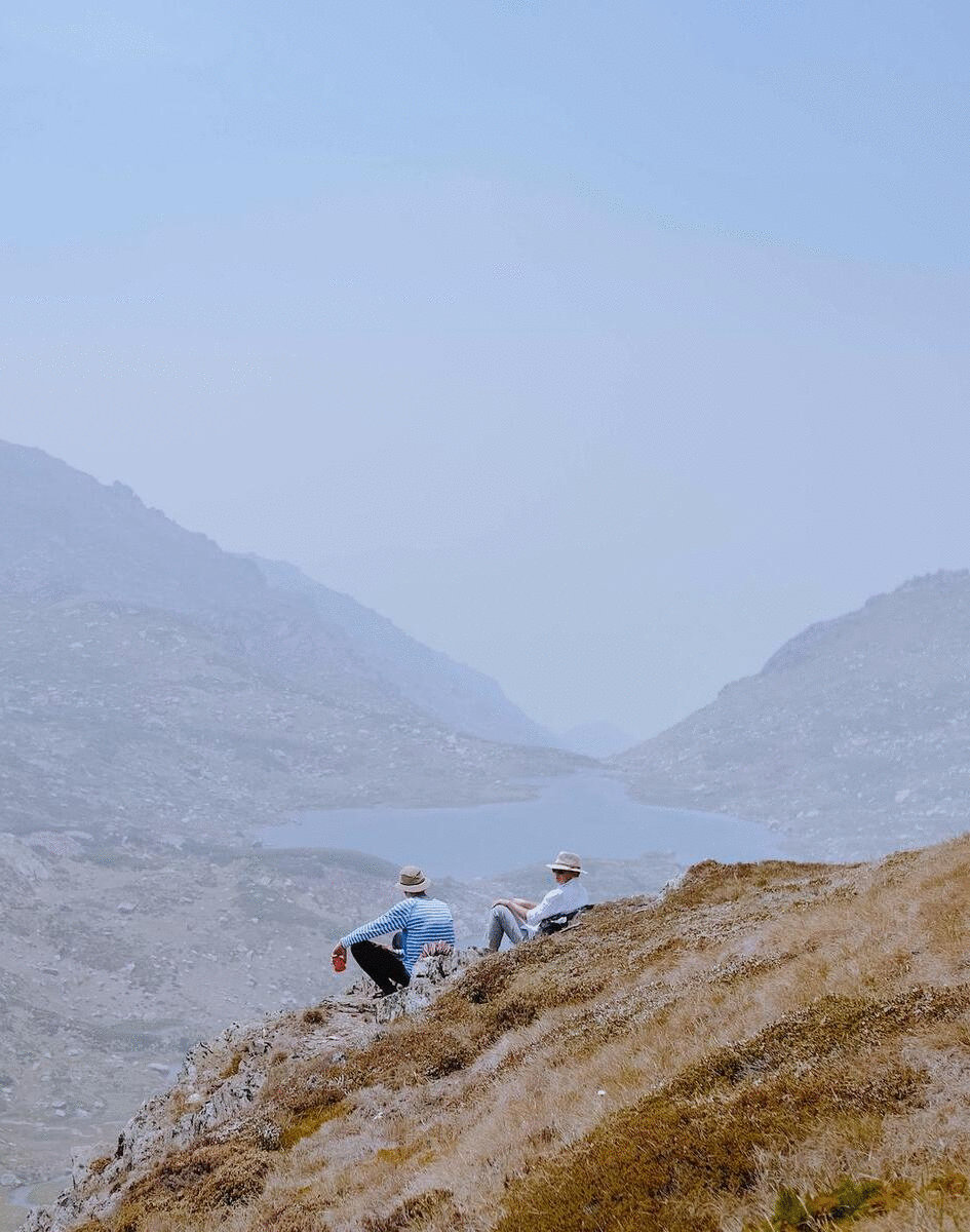 A couple sit atop a mountain and gaze out over a lake. 