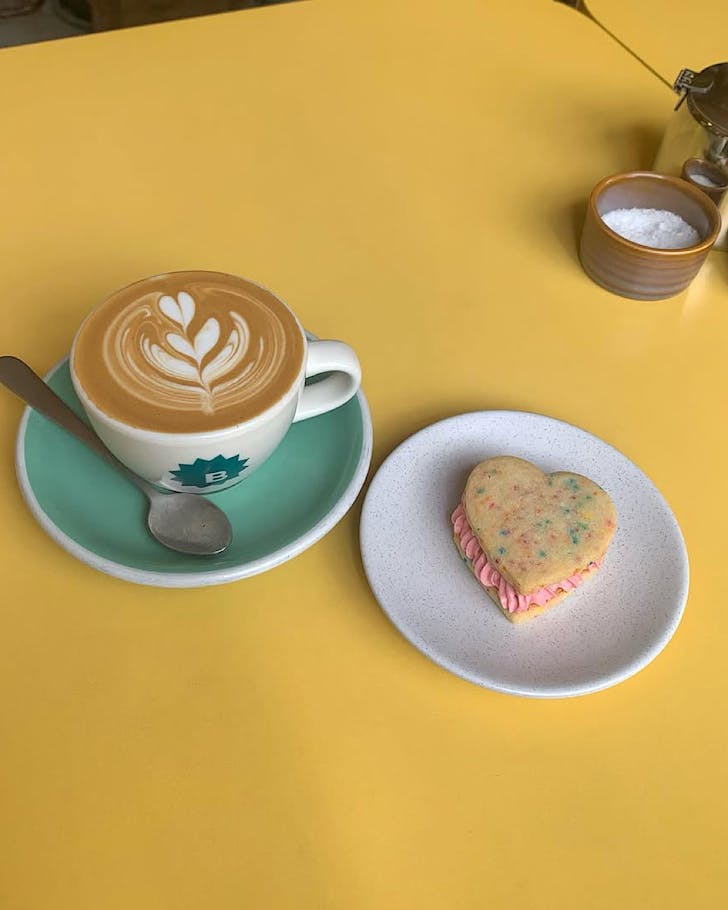 A cup of Bestie Cafe coffee sits next to a love heart-shaped cookie on a yellow table.