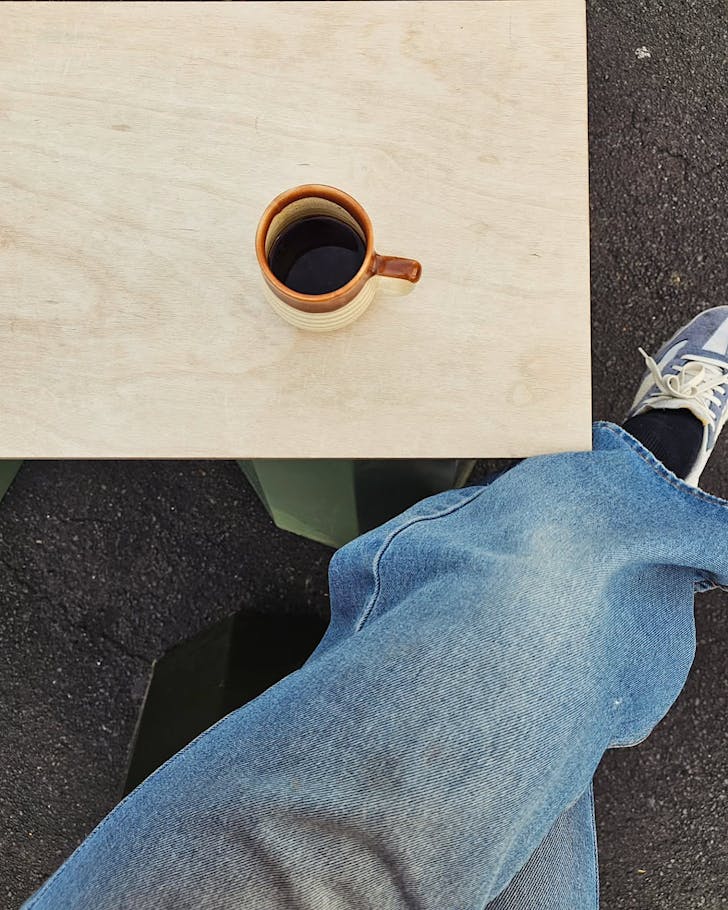 Someone in jeans and sneakers sits for a cup of ARK black coffee at a small table outside.
