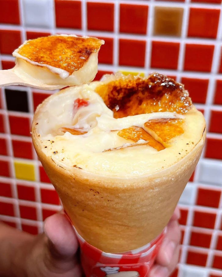 Close up image of Uncle Tetsu's Crepe Brulee, a crepe wrapped up like a cone with a thin layer of crispy cream brulee on top. The crispy layer in this photo has been slightly cracked as someone begins to scoop out the cheesecake inside.