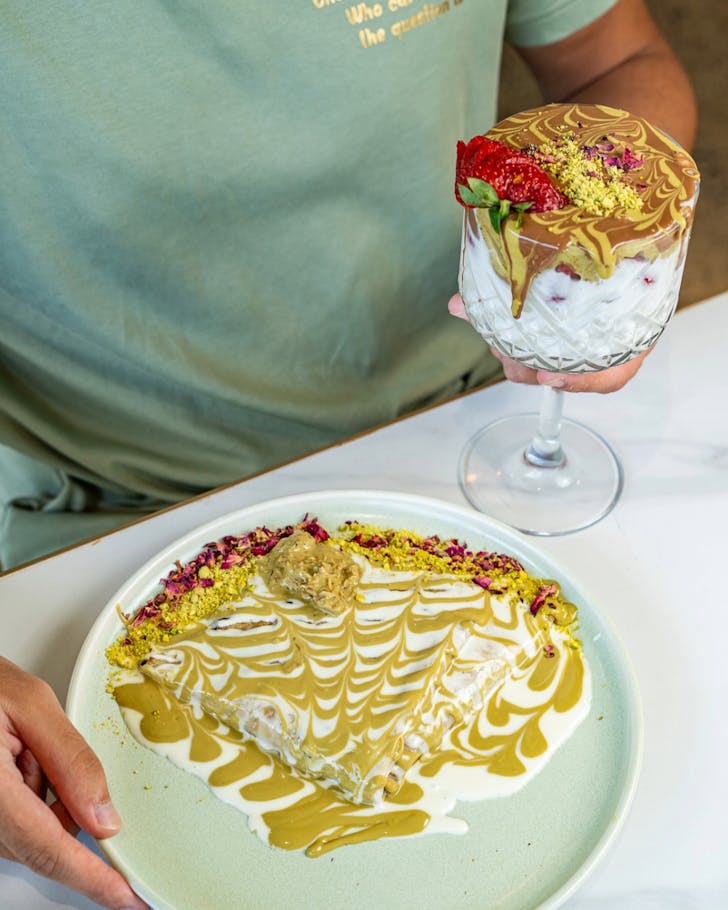 A view of a pistachio crepe and pistachio flavoured sweet drink on a table, sitting in front of a person who is about to eat it. The crepe and drink look creamy, rich, and colourful.
