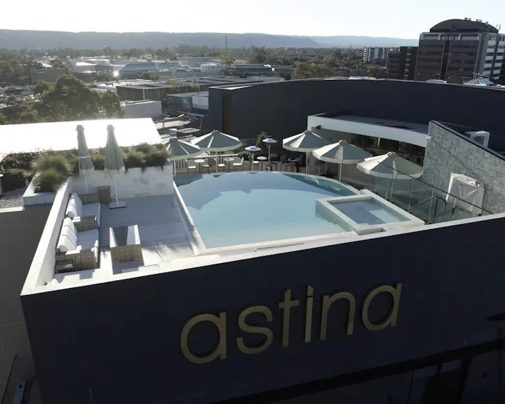 A wide birds eye view shot from the side of the Astina rooftop, where you can see Theo's Rooftop Bar in the daytime with a blue pool, umbrellas and dining tables. The background includes some buildings and mountains beneath the sky.