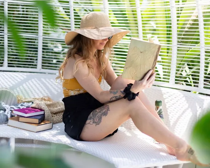 A mid shot of a lady sitting in swimwear on a pool chair, reading a book and with her face partially covered by a large sun hat. She is smiling gently, and there is greenery in the foreground and background, with daylight lighting filtering through the space she is in.