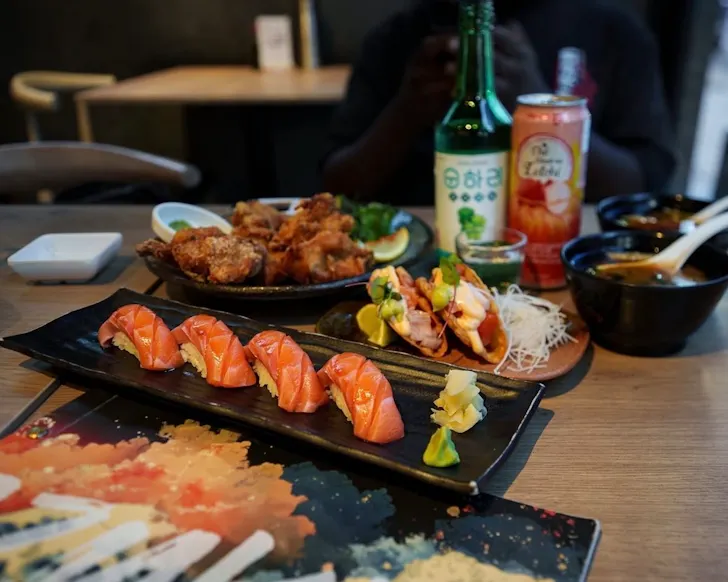 An close-up image of a small dining table in a restaurant where the focus is on a few dishes of sushimi, chicken, seafood tacos and some Japanese drinks that look aesthetically placed.